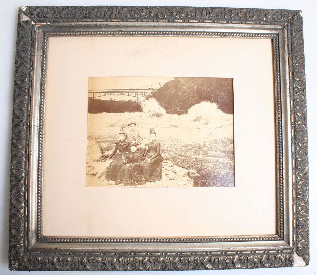 WOMEN AND MAN SEATED NEAR RAPIDS, TRAIN TRESTLE W/ TRAIN IN BACKGROUND. 1900.
