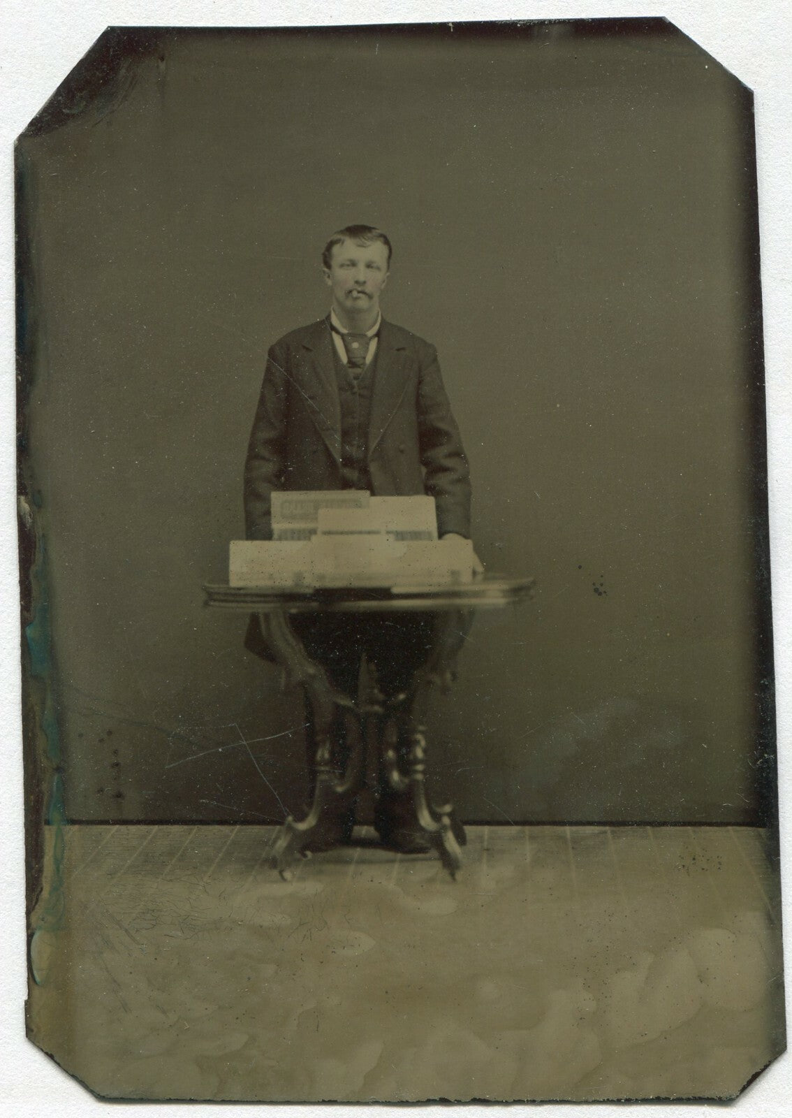 MAN POSED SMOKING CIGAR, CIGAR BOXES ON TABLE, TINTED TINTYPE.