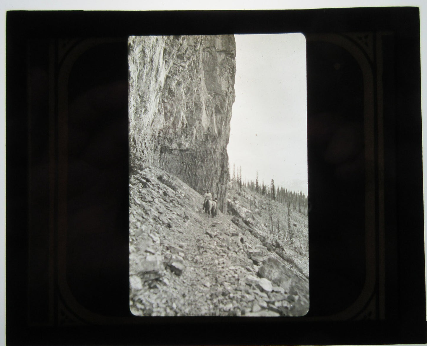COUPLE NAVIGATE ROCKY, MOUNTAIN SIDE ON HORSEBACK. LANTERN SLIDE.