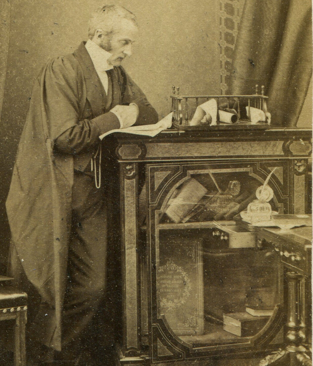 OLD MAN IN CAPE/LONG COAT AT STAGED DESK. ENGLAND.