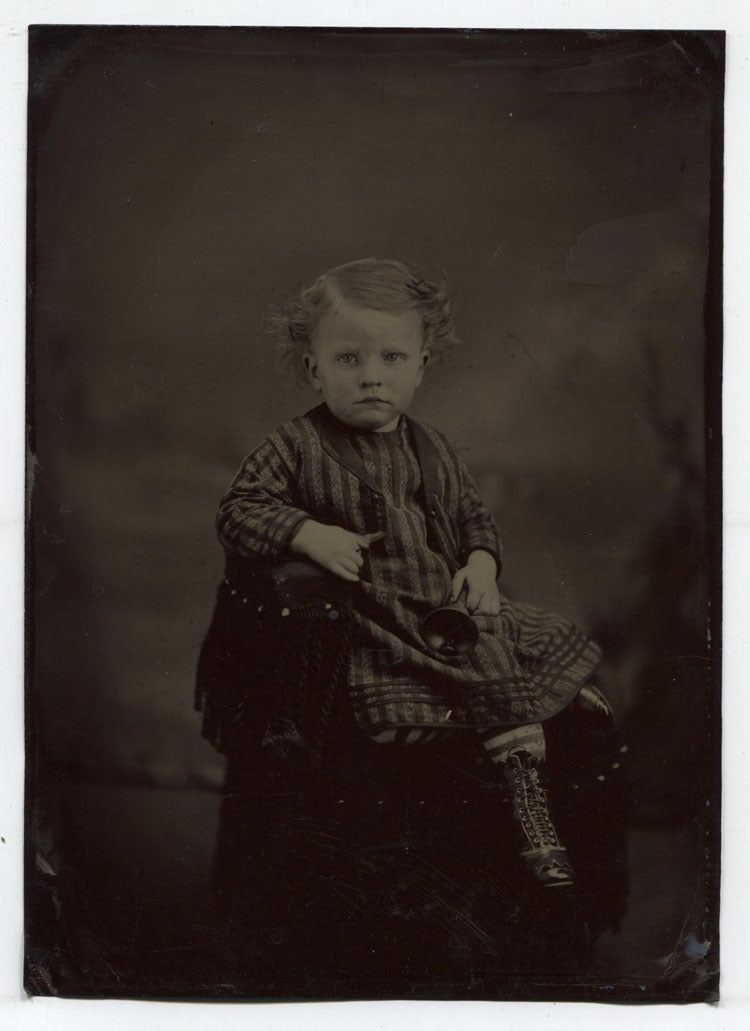 TINTYPE YOUNG CHILD IN STRIPED AND CHECK OUTFIT HOLDING A BELL. TINTED. UNHAPPY.