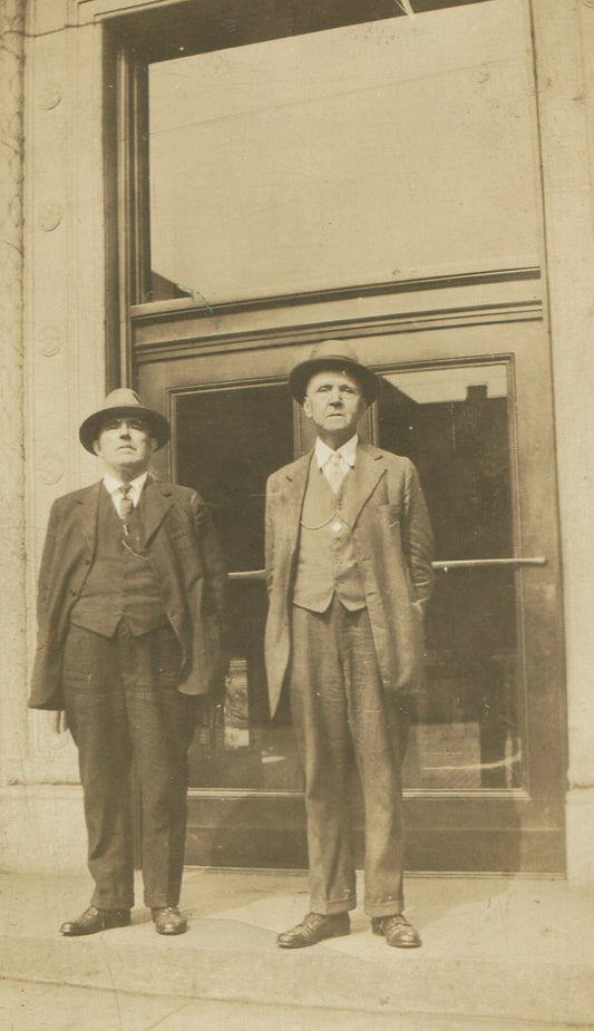 TWO MEN UNCOMFORTABLY POSED, STIFFLY STAND ON SIDEWALK, HUGE WINDOW. 1920.