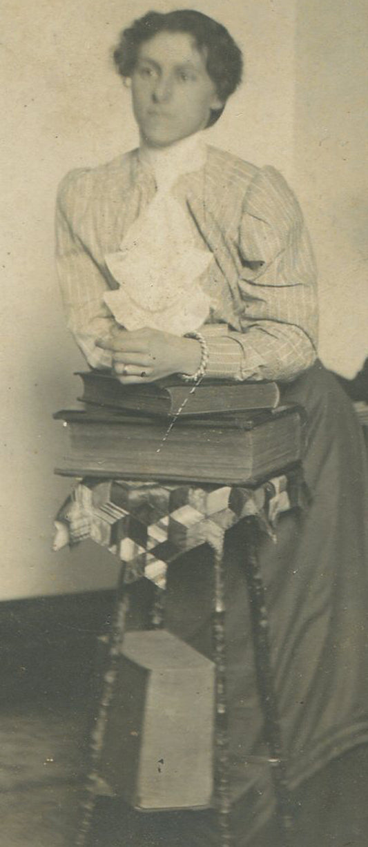WOMAN LEANING ON STACK OF BOOKS, INTERIOR. SILVER PRINT, CIRCA 1900.