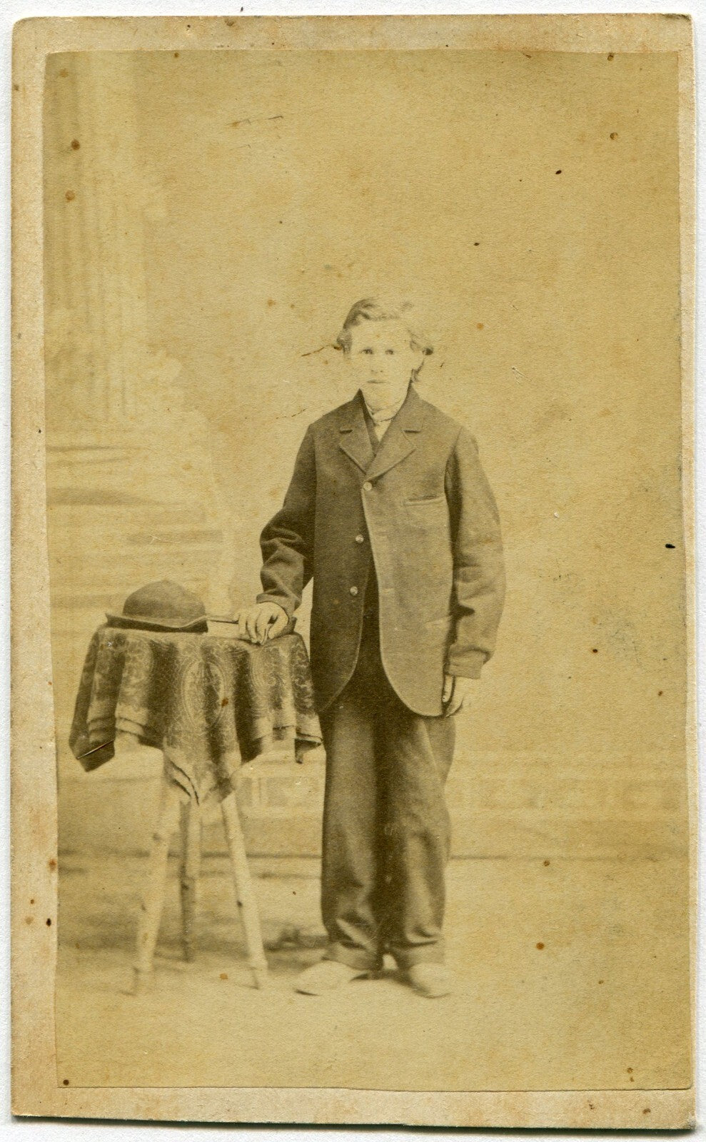 TALL GAUNT BOY IN BAGGY SUIT, HAT ON TABLE. CDV.