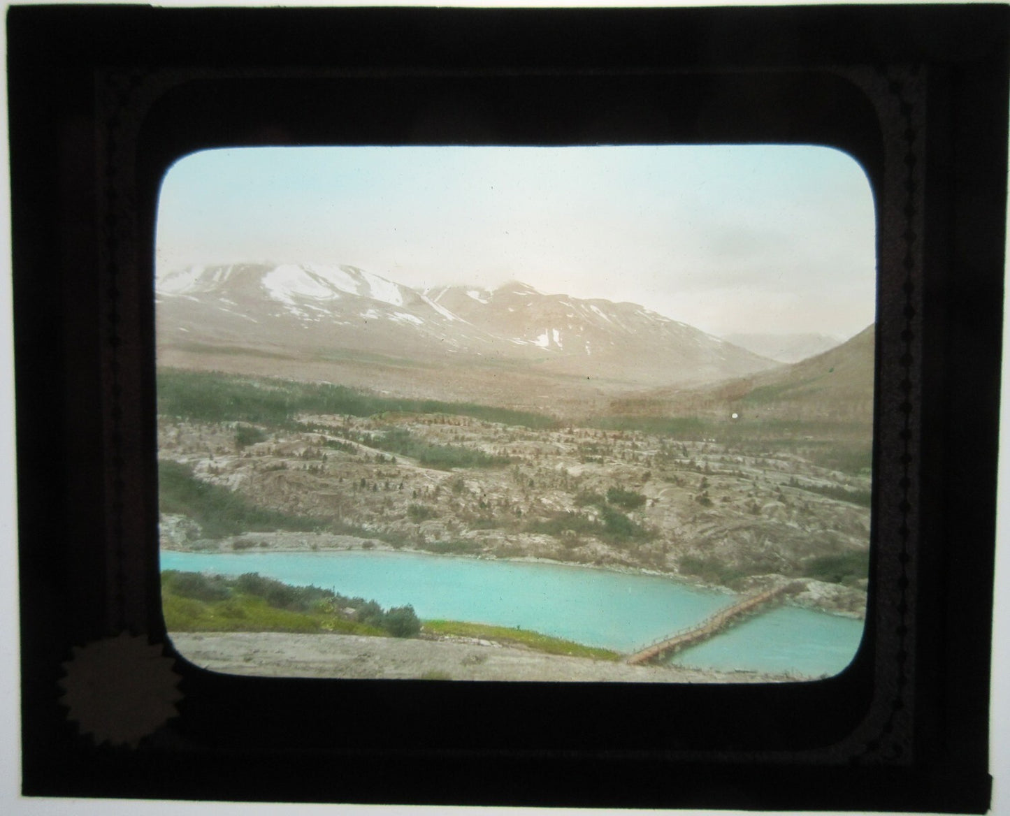 FOOT BRIDGE ON LAKE NEAR MOUNTAINS. HAND COLORED LANTERN SLIDE.