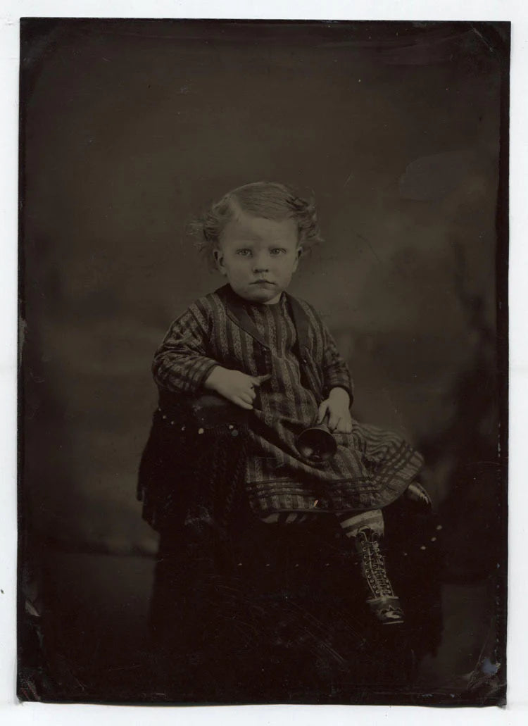 TINTYPE YOUNG CHILD IN STRIPED AND CHECK OUTFIT HOLDING A BELL. TINTED. UNHAPPY.