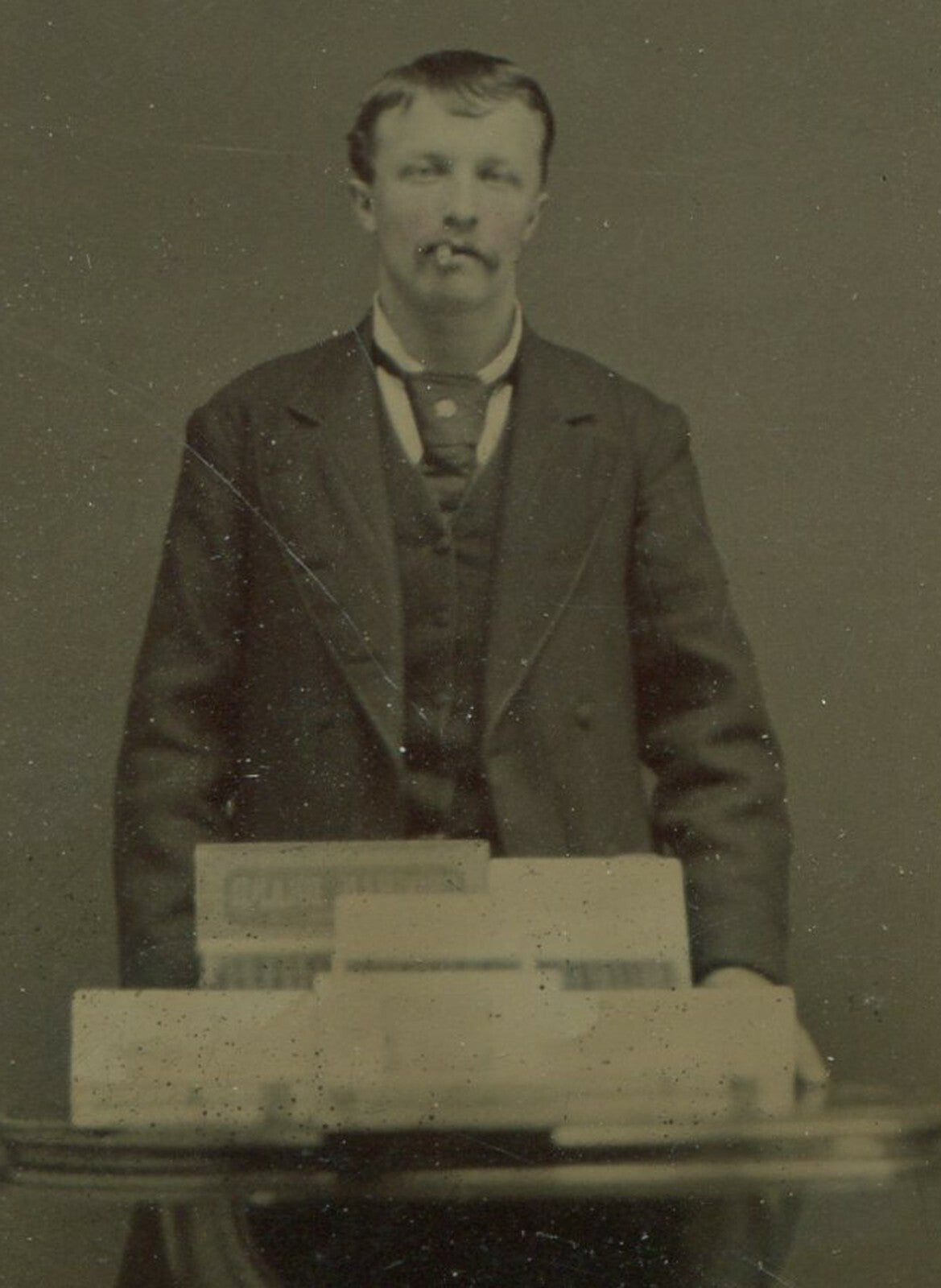 MAN POSED SMOKING CIGAR, CIGAR BOXES ON TABLE, TINTED TINTYPE.